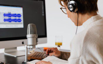 Imagem de uma mulher gravando em um estúdio em casa com microfone e uma tela de computador.