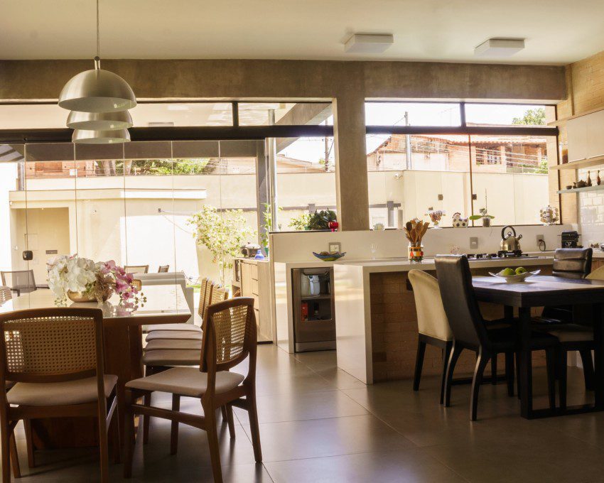 Área gourmet com mesa de jantar e espaço de cozinha ao fundo. Cadeiras, lustres e flores decoram o ambiente. Imagem disponível no Pexels