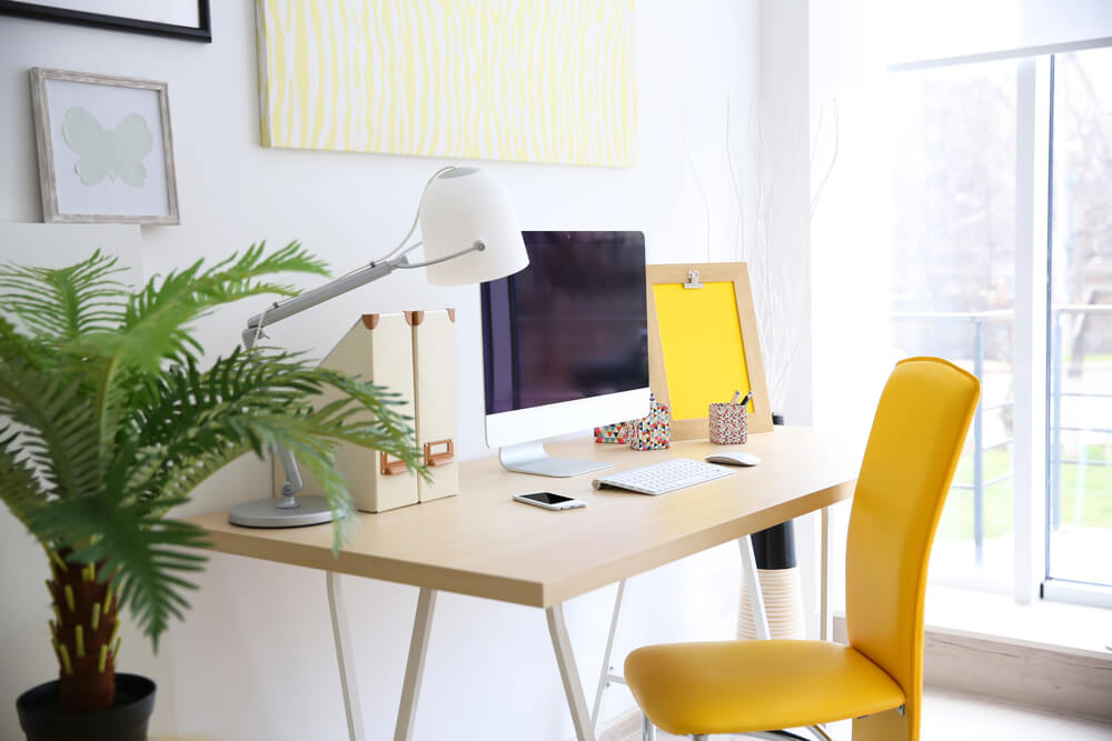Foto que ilustra matéria sobre decoração de escritório moderno mostra uma mesa com tampo de madeira clara com um computador e uma cadeira amarela em frente. 
