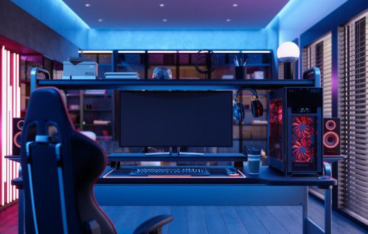 A foto mostra um exemplo de quarto gamer. Há uma poltrona gamer, uma mesa com monitor, teclado, mouse, fone de ouvido e CPU em cima dela. Há também uma iluminação especial no quarto, dando uma coloração em tom vermelho na lateral e azul neon no teto.