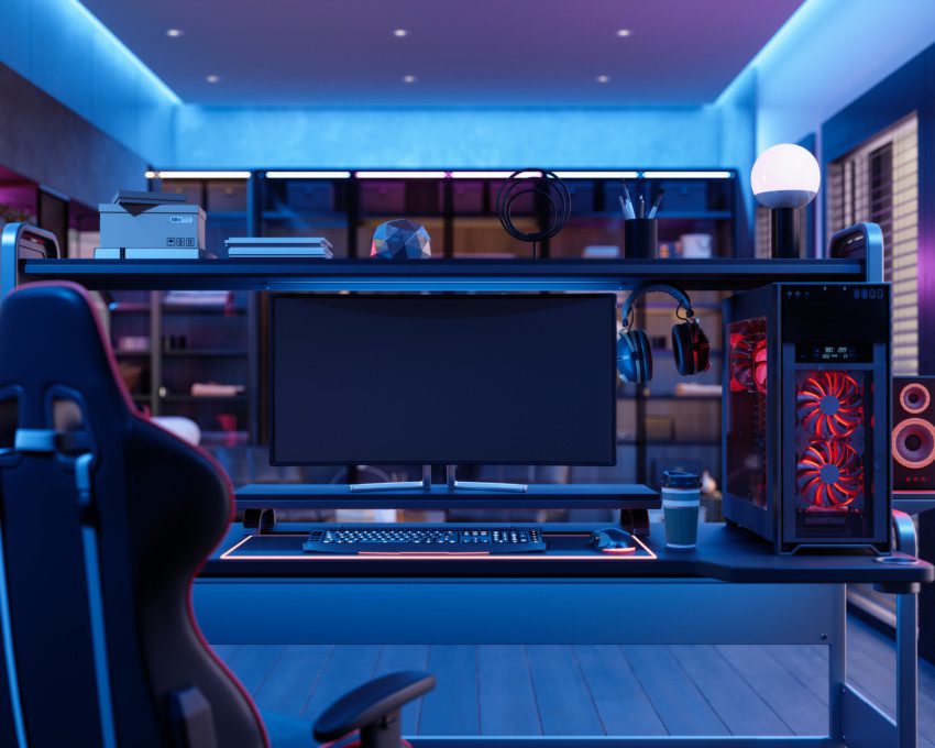 A foto mostra um exemplo de quarto gamer. Há uma poltrona gamer, uma mesa com monitor, teclado, mouse, fone de ouvido e CPU em cima dela. Há também uma iluminação especial no quarto, dando uma coloração em tom vermelho na lateral e azul neon no teto.