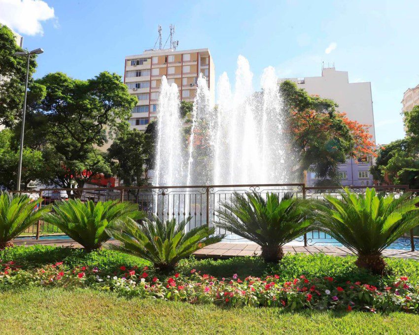 Foto que ilustra matéria sobre o que fazer em São José do Rio Preto mostra chafariz da Praça Rui Barbosa cercado por um gradeado, plantas, árvores e com prédios ao fundo em um dia de céu azul.