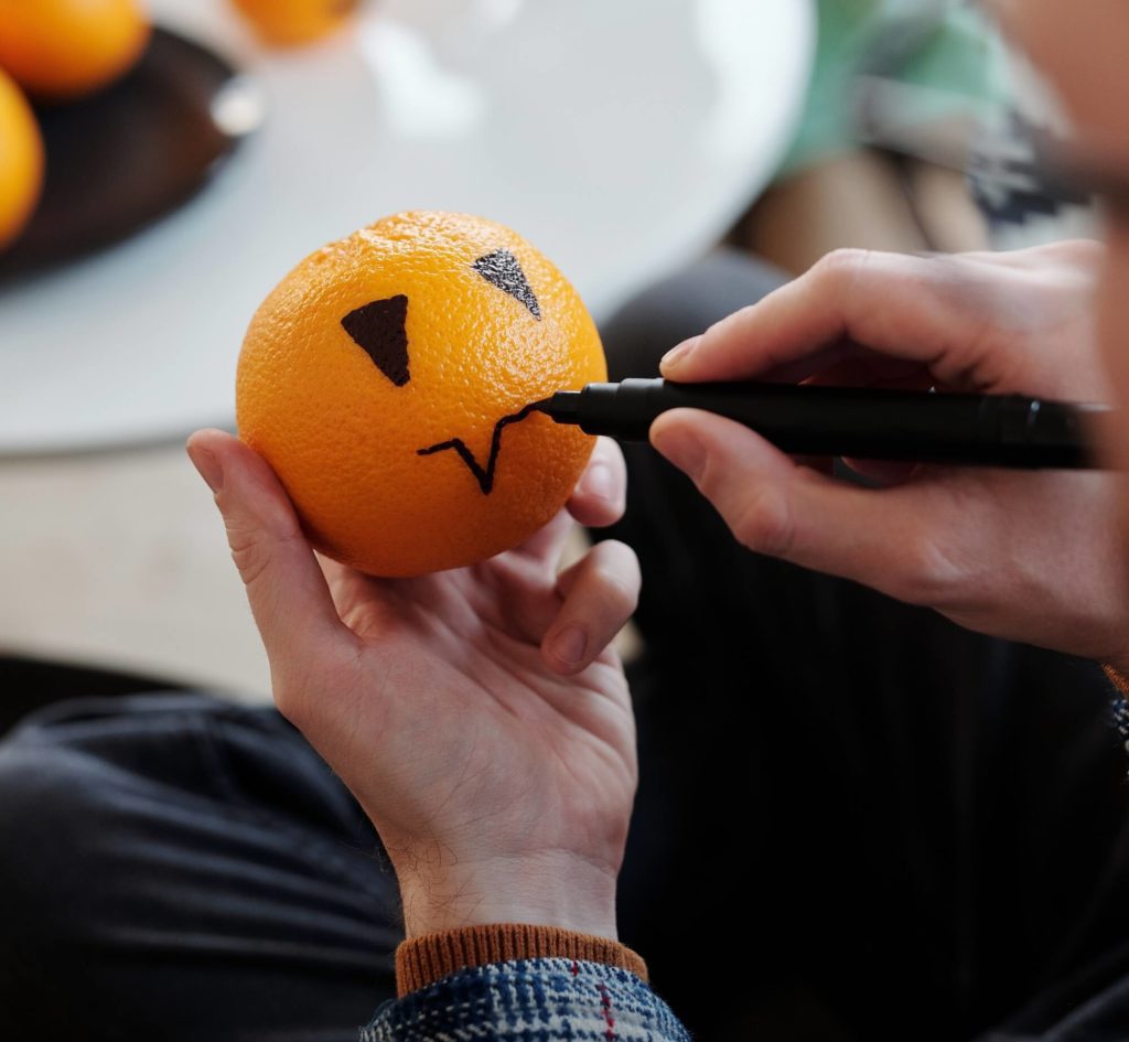 Foto que ilustra matéria sobre decoração de Halloween mostra uma mão segurando uma laranja enquanto a outra desenha nela uma carinha com caneta preta. 