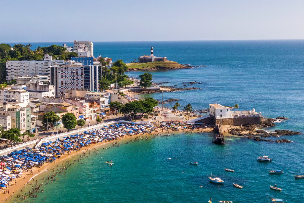 Imagem que ilustra matéria sobre o que fazer em Salvador mostra a vista da região da Barra em Salvador, vista de cima com prédios, guarda-sóis na areia e o mar. 