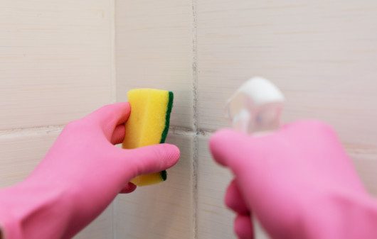 Imagem de uma pessoa limpando azulejo de um banheiro com esponja.