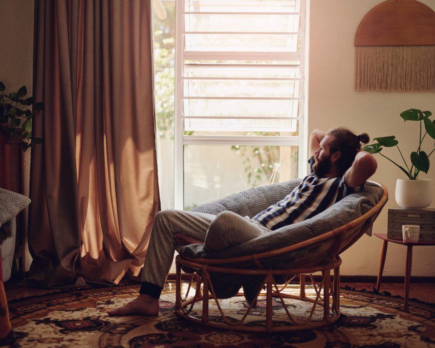 Imagem de uma homem sentado em uma poltrona de uma sala de estar com decoração em tons terrosos.