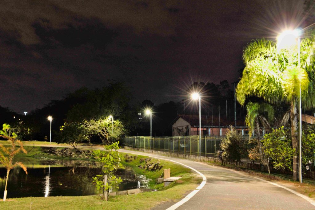 Foto que ilustra matéria sobre parque em Cotia mostra um trecho do Parque Teresa Maia à noite, com um caminho de concreto ao lado de um pequeno lago e com a iluminação acesa.