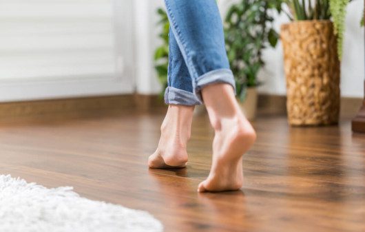 Foto que ilustra matéria sobre tipos de piso mostra o detalhe de um piso de madeira com os pés descalços de uma mulher andando sobre ele.