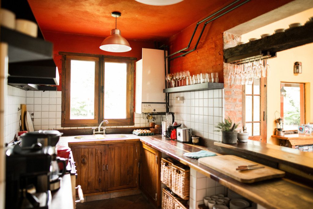 A imagem mostra o exemplo de uma cozinha rústica. Nela há um teto vermelho, uma luminária de cobre, uma pia com armário de madeira embutido. Há também diferentes utensílios como: cafeteira, tábua, facas, tostadeira, copos, taças, entre outros.