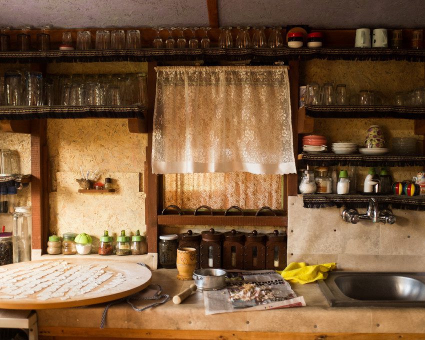 A foto mostra um exemplo de cozinha rústica em que há diferentes prateleiras de madeira ocupando toda a parede. Nelas tem utensílios como copos, xícaras, pratos, temperos, entre outros. Há também uma bancada de madeira com uma pia embutida.