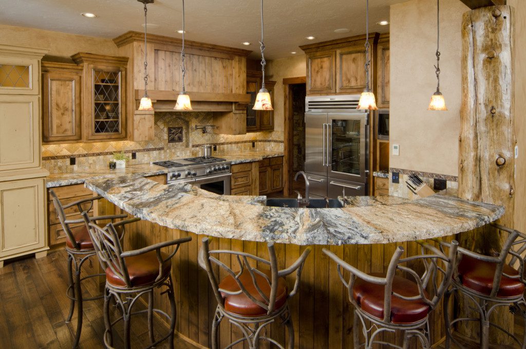 A imagem mostra uma cozinha americana rústica que tem uma bancada de mármore. Nela há também 5 banquetas de madeira, 5 luminárias de teto, uma geladeira e um fogão de inox. Além disso, tem um armários em toda a parede da cozinha.