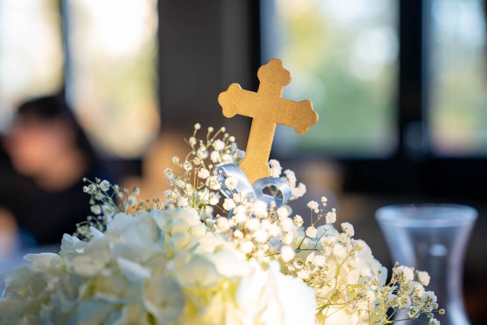 Foto que ilustra matéria sobre decoração para batizado mostra um centro de mesa com um arranjo de flores brancas e uma cruz dourada. 