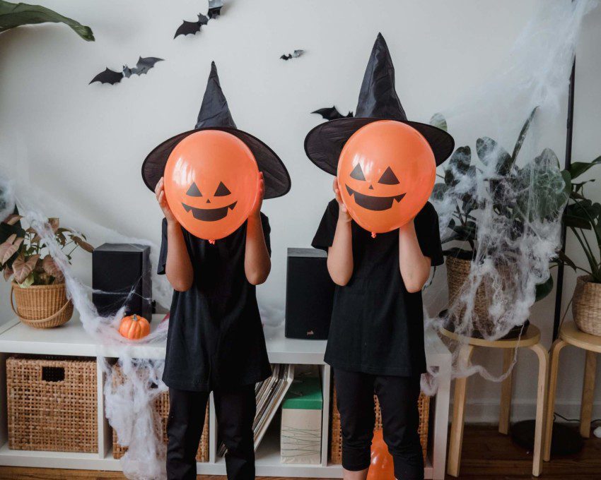 Foto que ilustra matéria sobre decoração de Halloween mostra duas crianças vestidas de preto, com chapéus de bruxa preto e com balões na cor laranja imitando abóboras com carinhas em frente a seus rostos. Na parede atrás deles, há pequenos morcegos feitos de papel pendurados.