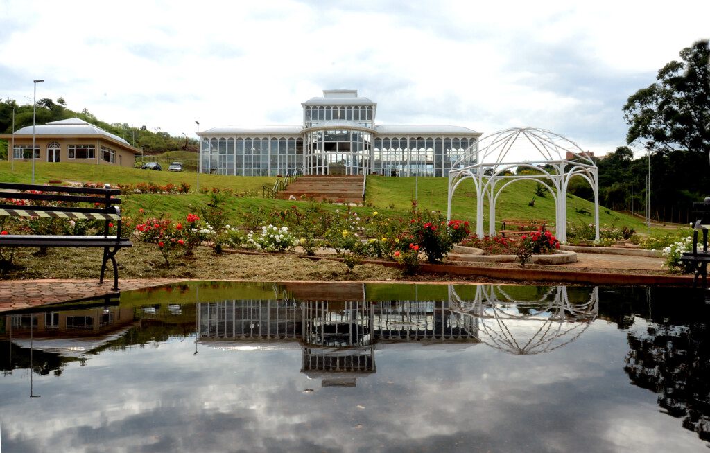 Imagem panorâmica com vista para o Palacete de Cristal do Jardim Botânico de Sorocaba cercado por flores e um espelho d'água 
