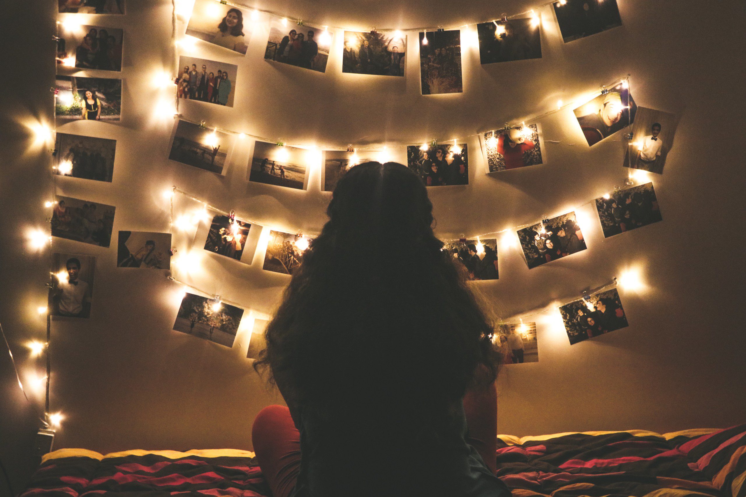 Imagem de uma mulher sentada na cama observando um varal de fotos com luz. 