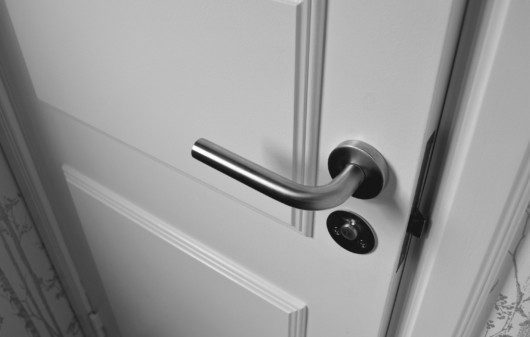 Imagem que ilustra matéria sobre tipos de fechadura mostra uma fechadura de alumínio em uma porta branca que está fechada.
