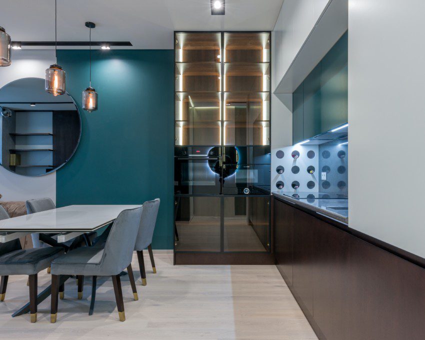 Foto de uma sala de jantar moderna com boa circulação. Na foto há uma mesa retangular com 4 cadeiras, uma estante em vidro, um espelho redondo e 3 lustres modernos no teto. Há também uma parede pintada em azul petróleo.