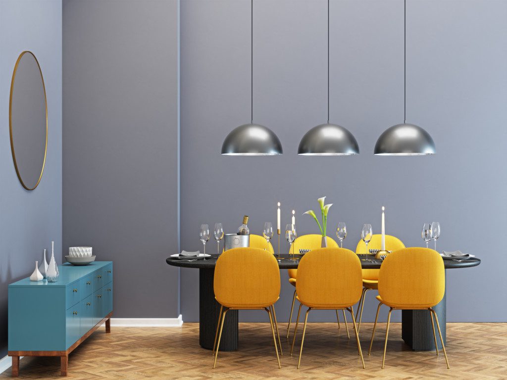 A foto mostra um exemplo de sala de jantar moderna com buffet. Há na imagem uma mesa oval com 6 cadeiras, 3 lustres redondos pendurados, um espelho redondo na parede e um buffet azul.