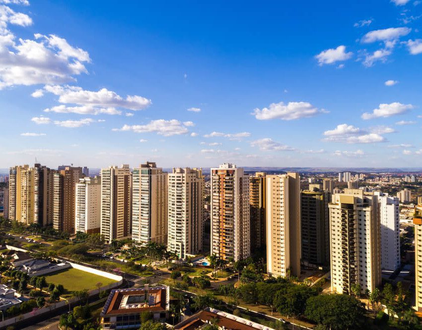 Foto que ilustra matéria sobre bairros de Ribeirão Preto mostra uma visão do alto da cidade, com diversos prédios grandes no centro da imagem, áreas arborizadas no canto inferior esquerdo e um céu azul com poucas nuvens brancas de um dia claro.