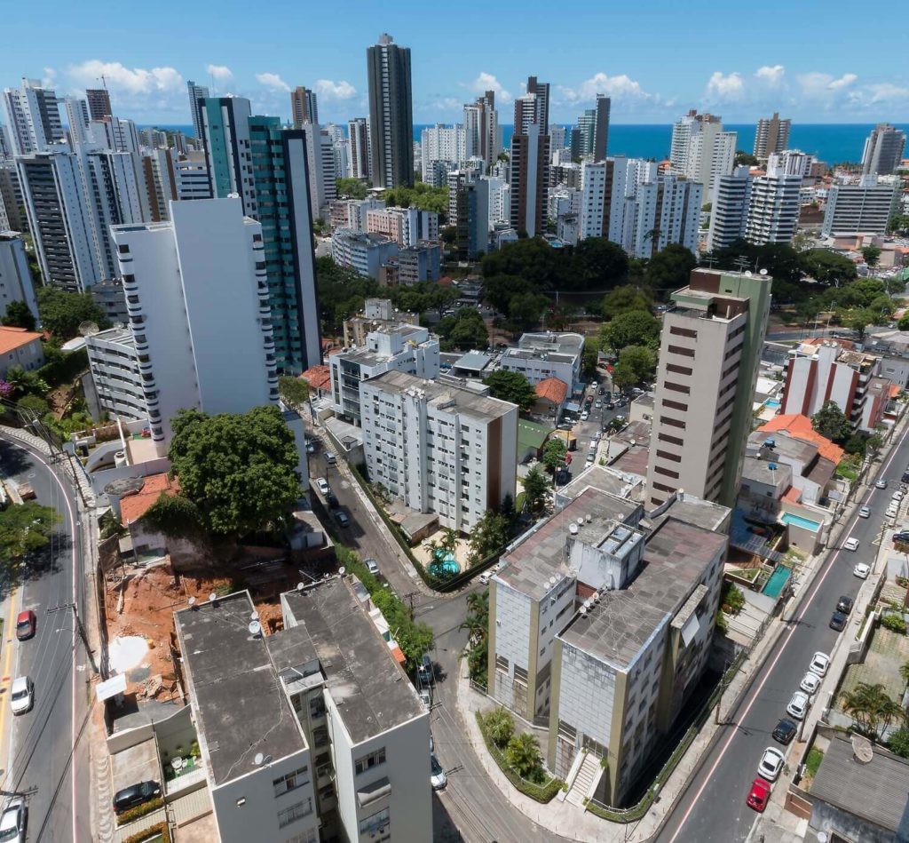 Foto que ilustra matéria sobre bairros mais seguros de Salvador mostra prédios altos que cercam uma área arborizada e que têm ao fundo o mar e o céu azuis. 