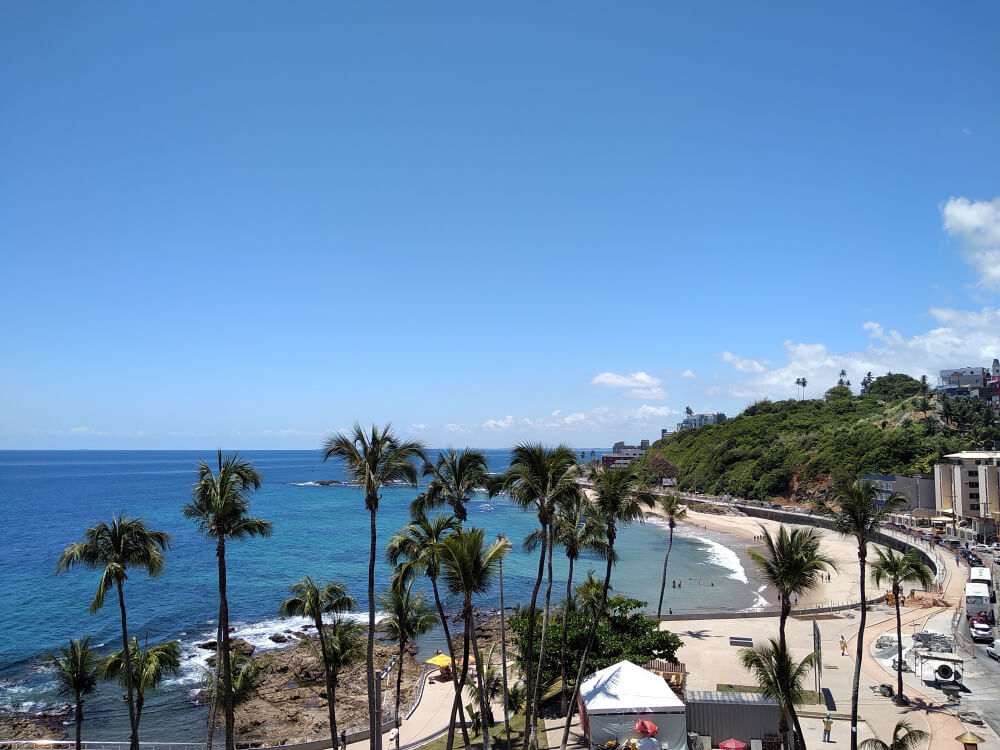 Foto que ilustra matéria sobre bairros mais seguros de Salvador mostra uma vista aérea de uma praia, a de Ondina, em Salvador, em que o mar azul aparece à esquerda e o calçadão aparece à direita.