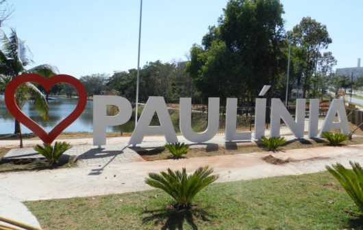 Foto que ilustra matéria sobre como chegar em Paulínia mostra uma imagem de um letreiro em um parque com um coração vermelho e o nome da cidade escrito em branco. Atrás, aparecem um lago, árvores e um céu azul claro.