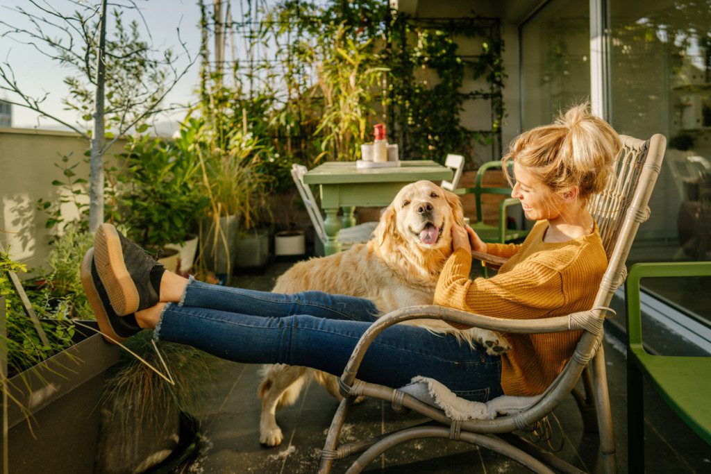 Imagem que ilustra matéria sobre varanda aconchegante mostra uma mulher fazendo carinho em um cachorro em uma varanda cheio de folhagens. 