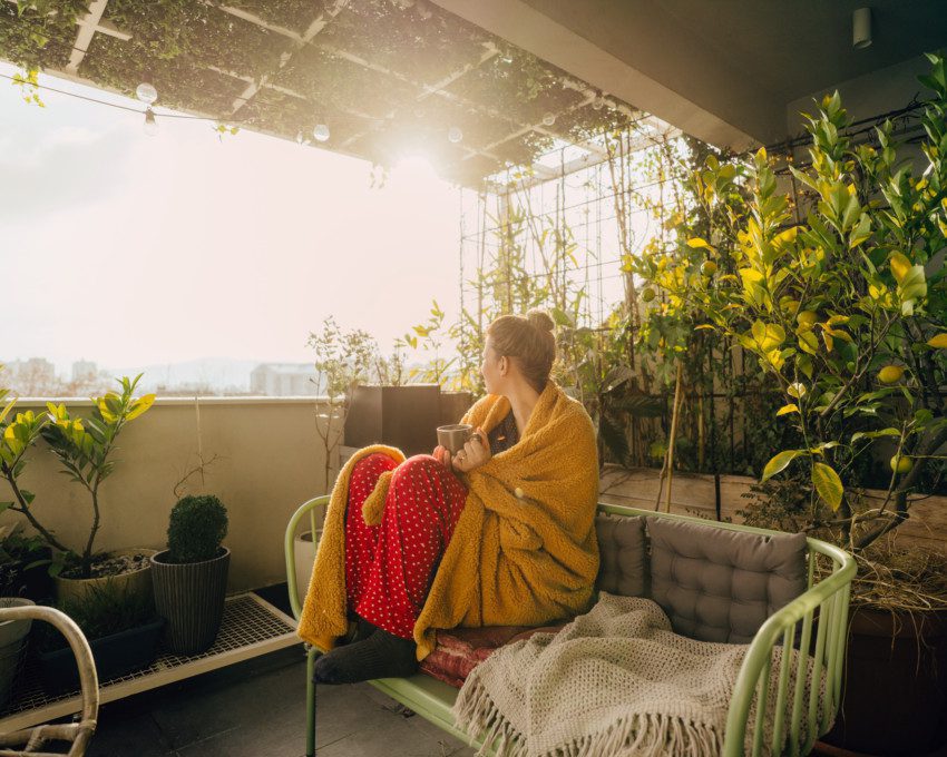 Foto de uma varanda com uma mulher sentada no sofá. Ao redor dela há plantas de diferentes tamanhos.