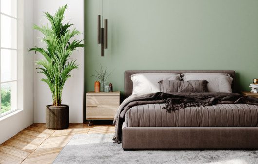Imagem de um quarto com cama de casal encostada em uma parede verde, mesa de cabeceira e um vaso de planta.