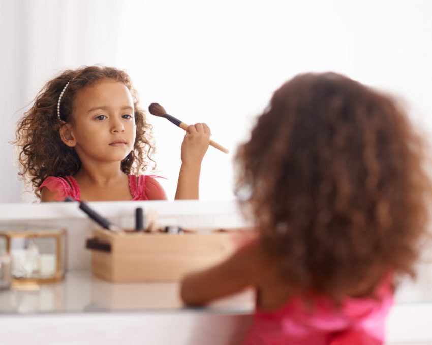 Imagem de uma menina segurando um pincel de maquiagem sentada de frente para uma penteadeira.