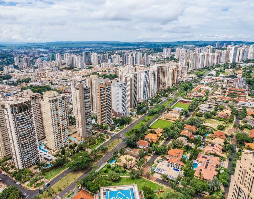 Foto que ilustra matéria sobre morar em Ribeirão Preto mostra uma visão do alto da Avenida Professor João Fiúza, a mais importante da cidade, com muitos prédios altos à esquerda e casas em zonas arborizadas à direita.