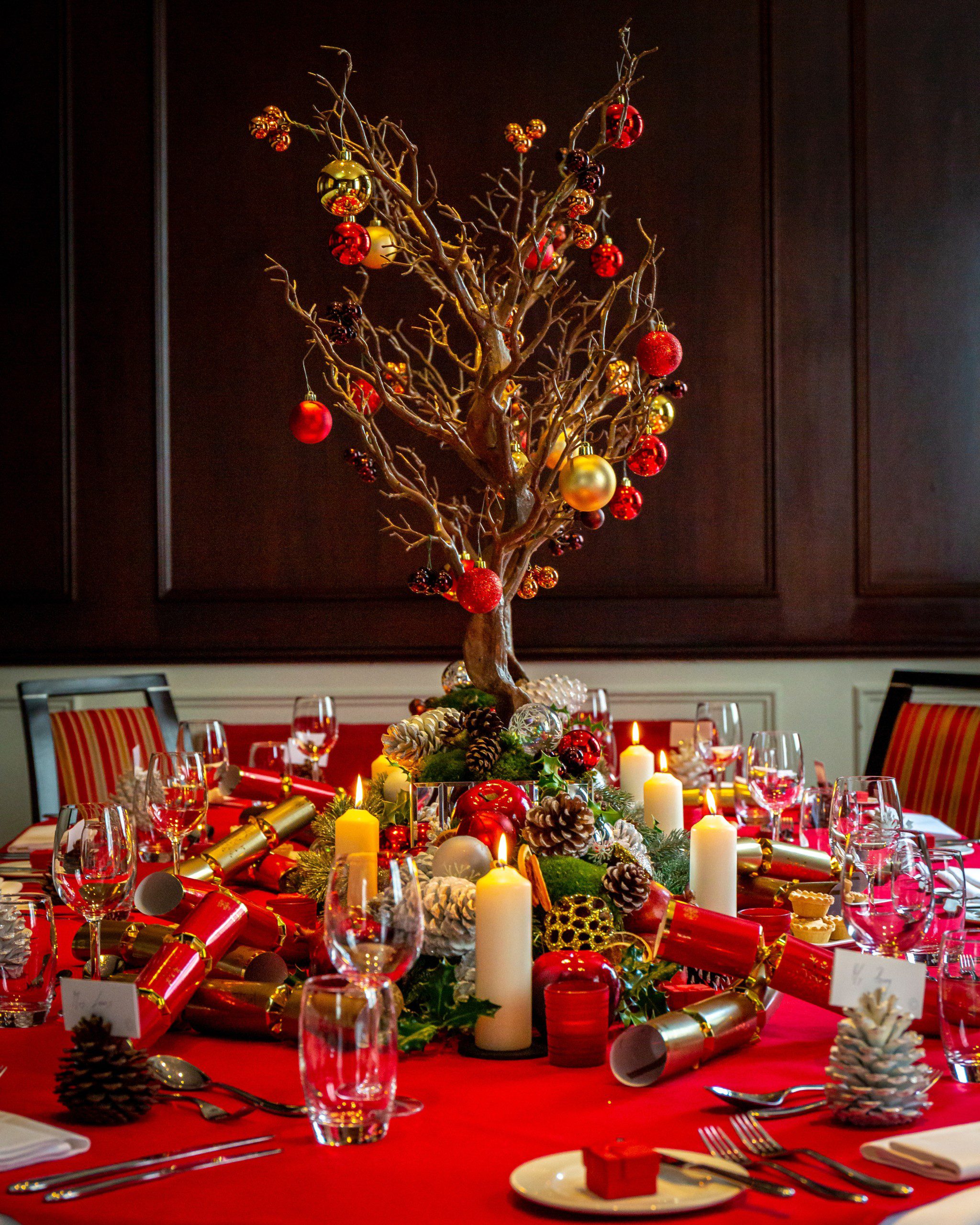 Mesa de Natal com toalha vermelha e uma árvore com galhos centro como enfeite de centro.