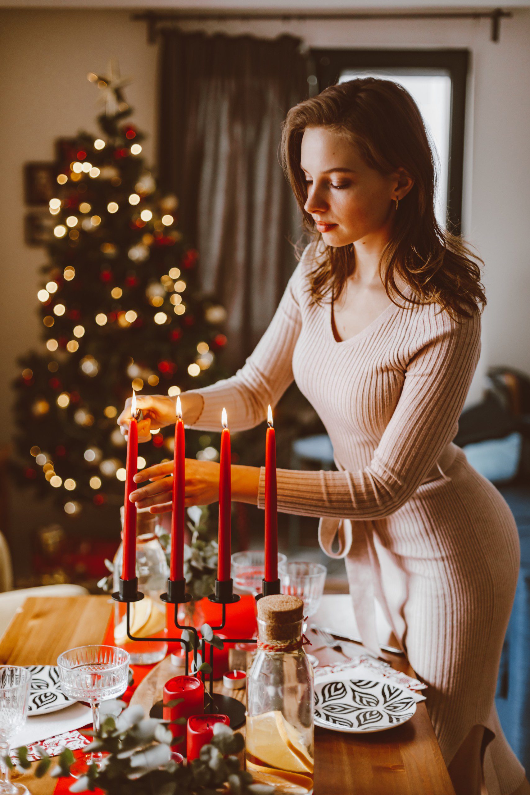 Uma mulher acendendo velas vermelhas em uma mesa natalina com uma árvore de fundo.