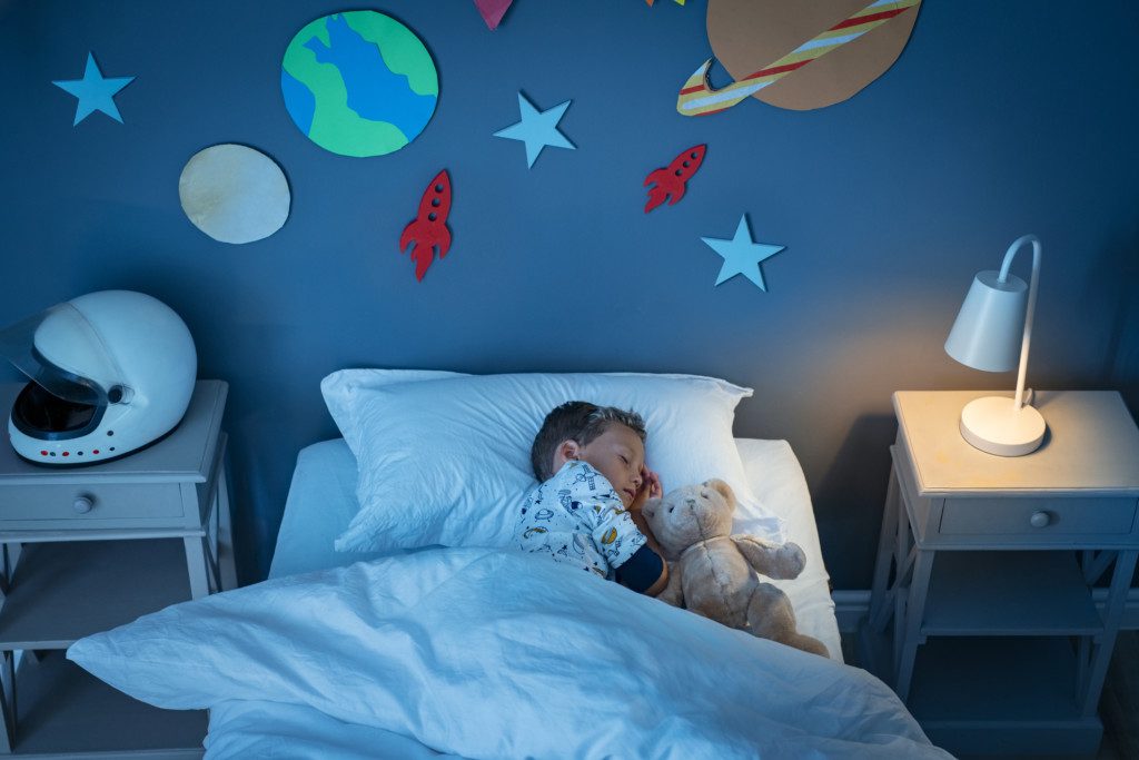 Foto de uma criança dormindo na cama. Em cima dela, na parede, há diferentes desenhos do espaço como planetas, estrelas e foguetes. Tem também na imagem duas mesas de cabeceiras, uma delas com uma luminária e outra com um capacete de astronauta.