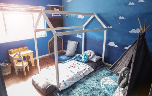 Imagem de um quarto com paredes pintadas de azul. Nelas há nuvens imitando o céu. Nele há também uma cama, uma cabana, almofadas de bichos e uma escrivaninha pequena com cadeira para criança.