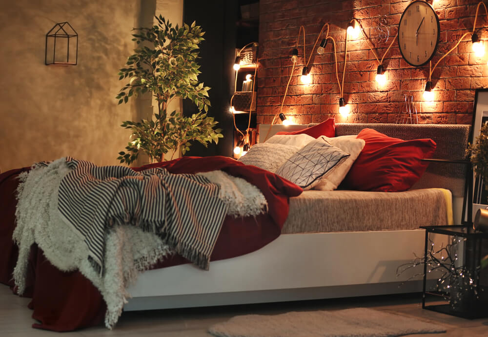 Foto que ilustra matéria sobre como decorar o quarto gastando pouco mostra uma cama com sua cabeceira encostada em uma parede de tijolos onde há um varal de luzes pendurado e iluminando o ambiente
