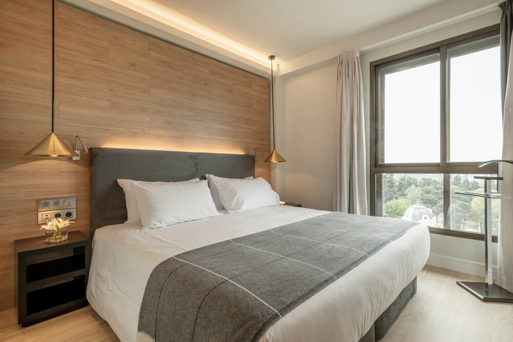 Foto que ilustra matéria sobre como decorar o quarto gastando pouco mostra uma cama com duas mesas laterais com luminárias pendentes e uma cabeceira com luz indireta atrás