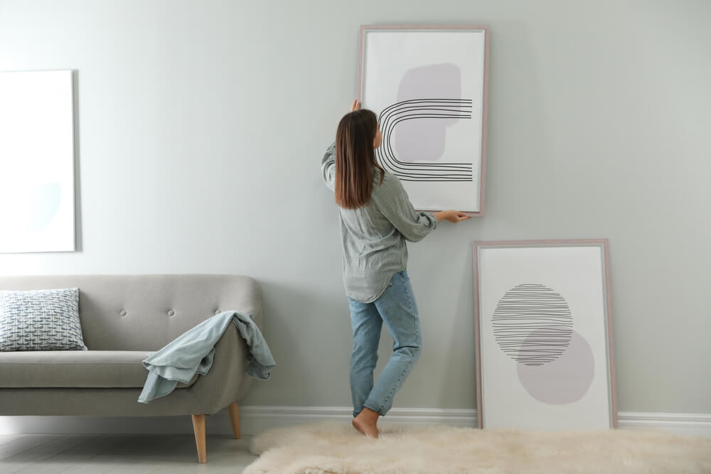 Foto que ilustra matéria sobre como decorar a sala gastando pouco mostra uma mulher de costas pendurando quadros em uma parede