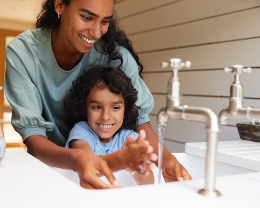 Foto que ilustra matéria sobre consumo consciente de água mostra uma torneira aberta onde aparece em detalhe duas mãos de uma pessoa que se lava.