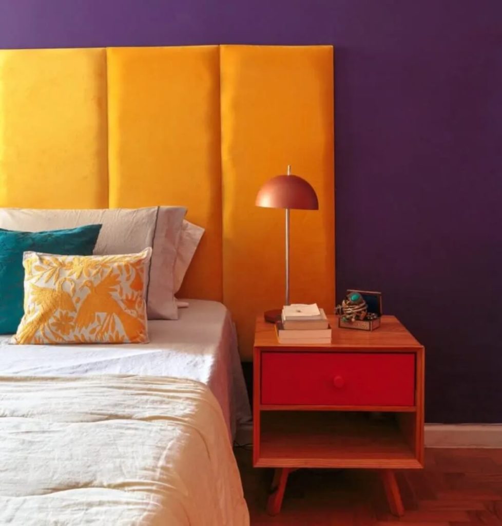 Foto que ilustra matéria sobre como decorar o quarto gastando pouco mostra uma parte de uma cama, com destaque para a cabeceira amarela, a parede roxa e a mesinha de cabeceira de madeira com gaveta vermelha