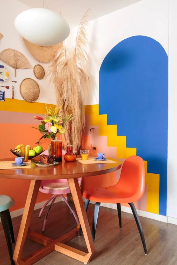 Foto que ilustra matéria sobre pintura setorizada mostra uma sala de jantar com pinturas diferentes nas paredes, como um arco azul sobreposto por lustras amarela, bege e laranja em formato de escada.