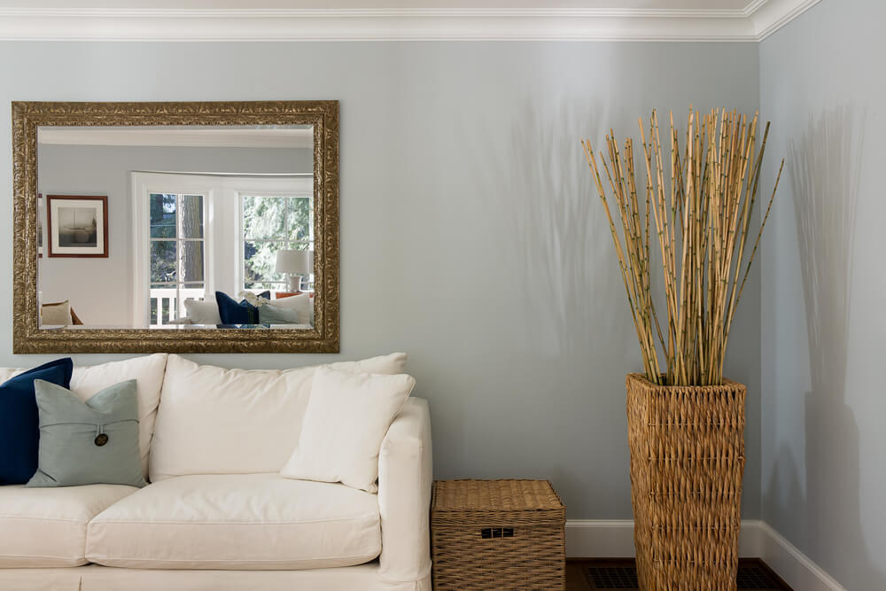 Foto que ilustra matéria sobre como decorar a sala gastando pouco mostra um pedaço de uma sala de estar com um sofá com um grande espelho acima dele na parede. Ao lado, um cesto e um vaso de vime com varetas de bambu