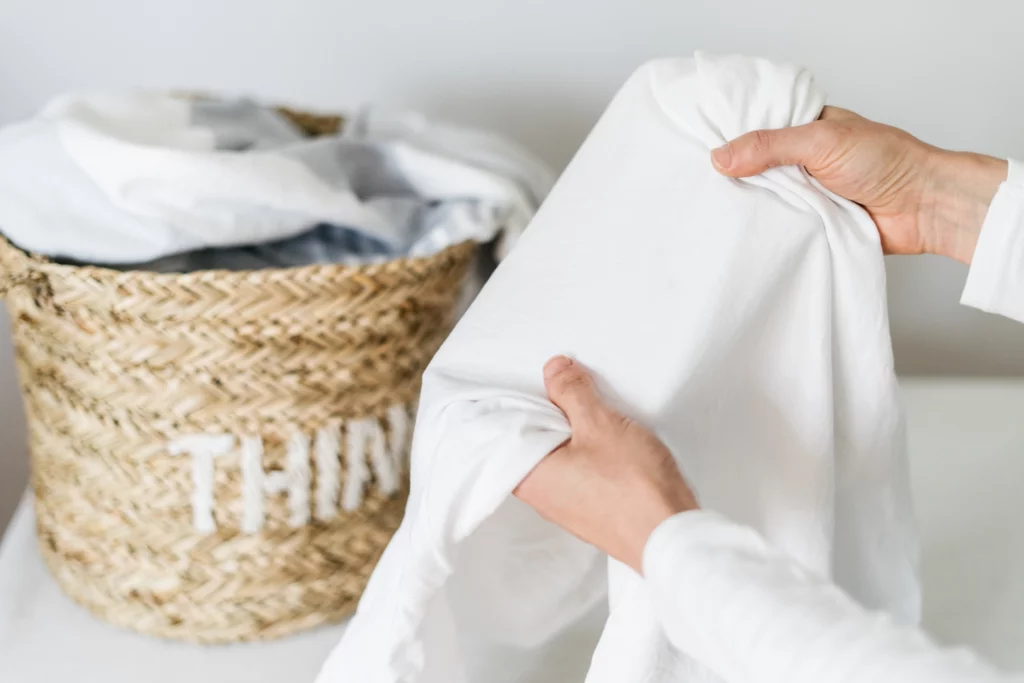Imagem de uma pessoa segurando roupa de cama limpa dentro de casa em frente a um cesto com mais roupas de cama, todas brancas, para ilustrar matéria sobre como clarear roupa branca