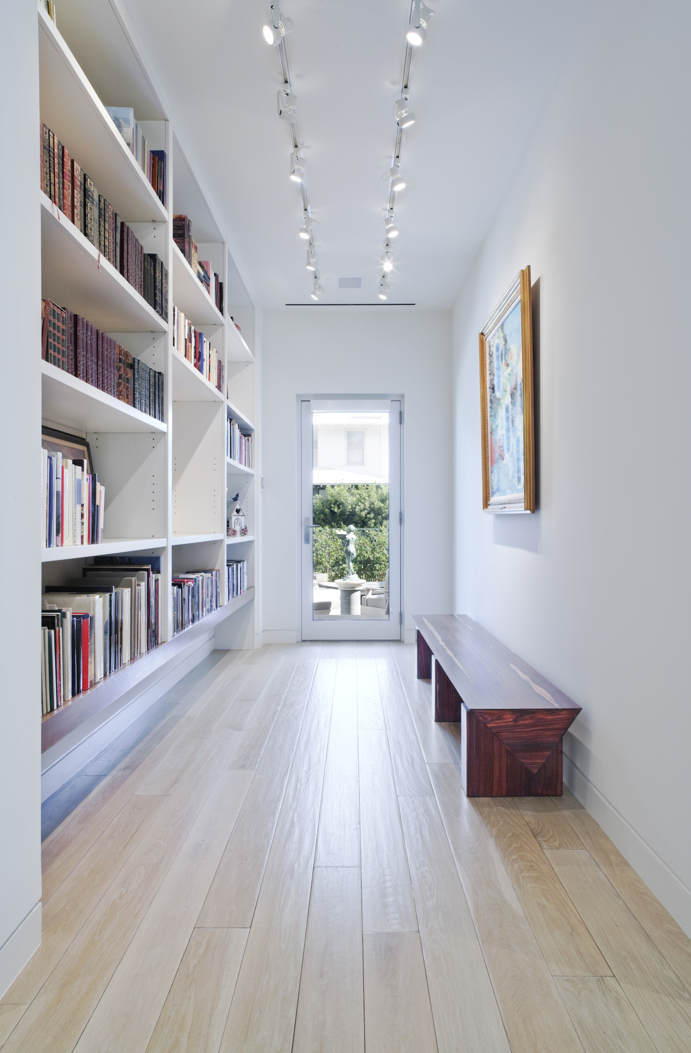 Foto de um corredor longo que tem de um lado da parede uma estante de livros e do outro uma banqueta. Há também na imagem um quadro perto da banqueta.