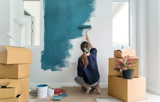 Imagem de uma mulher pintando uma parede branca com tinta azul e com caixa de papelão em volta.