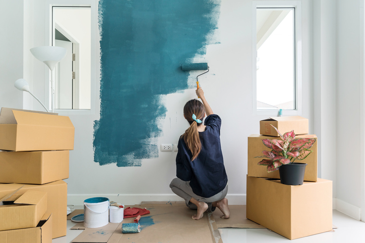 Imagem de uma mulher pintando uma parede branca com tinta azul e com caixa de papelão em volta.