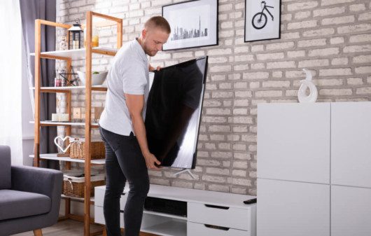 Foto que ilustra matéria sobre quanto custa mobiliar um apartamento mostra um homem segurando uma grande TV de LED para posicioná-la sobre um rack branco em uma sala de estar.