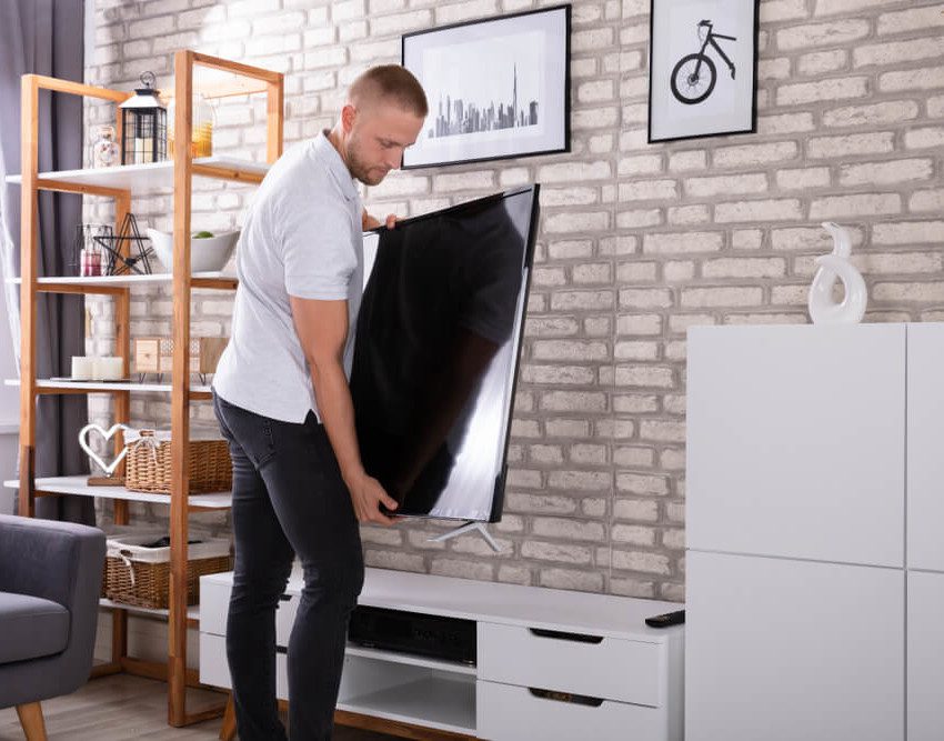 Foto que ilustra matéria sobre quanto custa mobiliar um apartamento mostra um homem segurando uma grande TV de LED para posicioná-la sobre um rack branco em uma sala de estar.