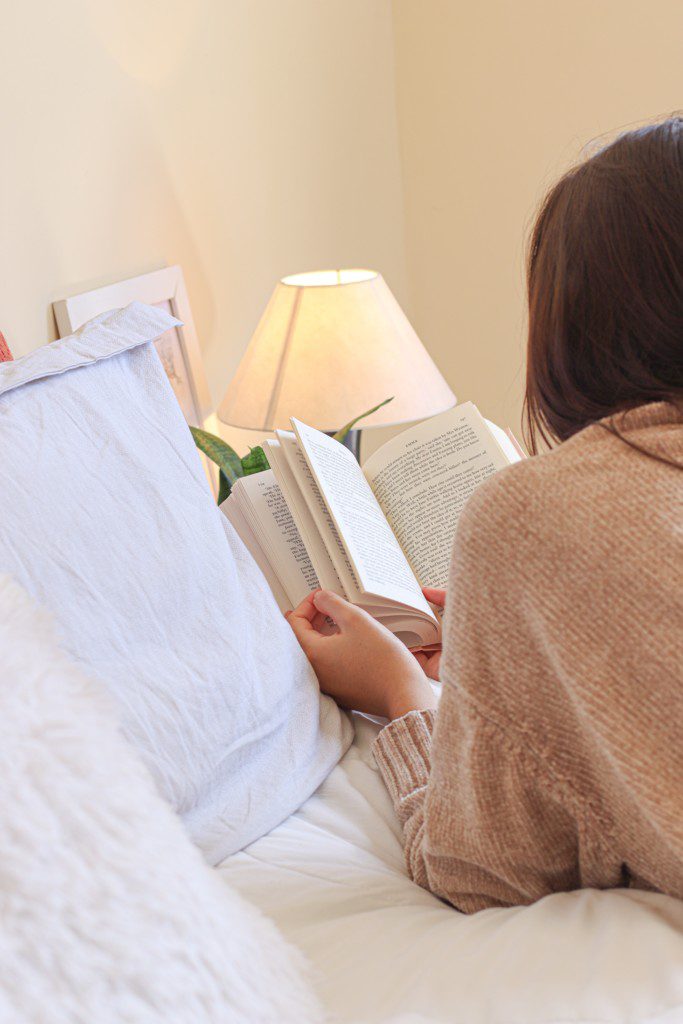 Mulher lê um livro em quarto de visita ao lado de um abajur. Imagem disponível em Pexels.