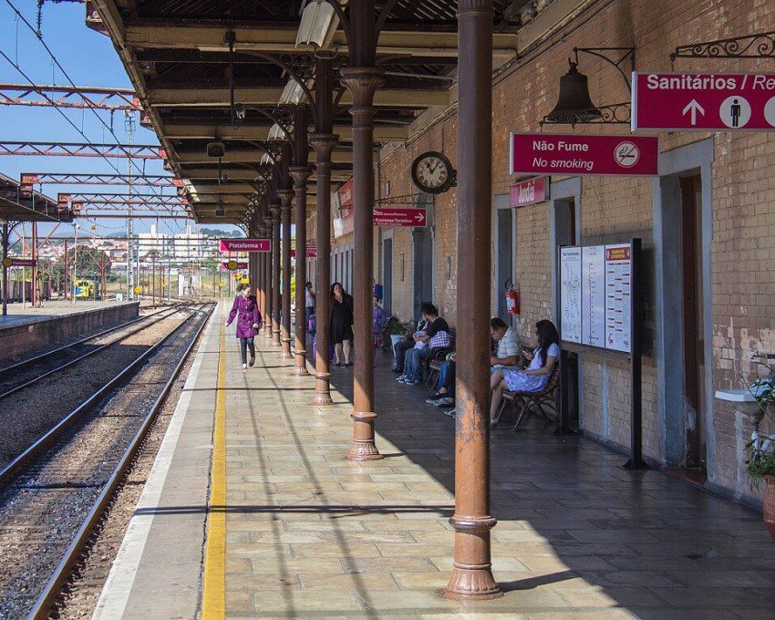 Foto que ilustra matéria sobre a Estação Jundiaí mostra a plataforma da estação de trem em um dia de céu azul, com pessoas sentadas aguardando a chegada do veículo à direita da imagem e os trilhos vazios à esquerda.